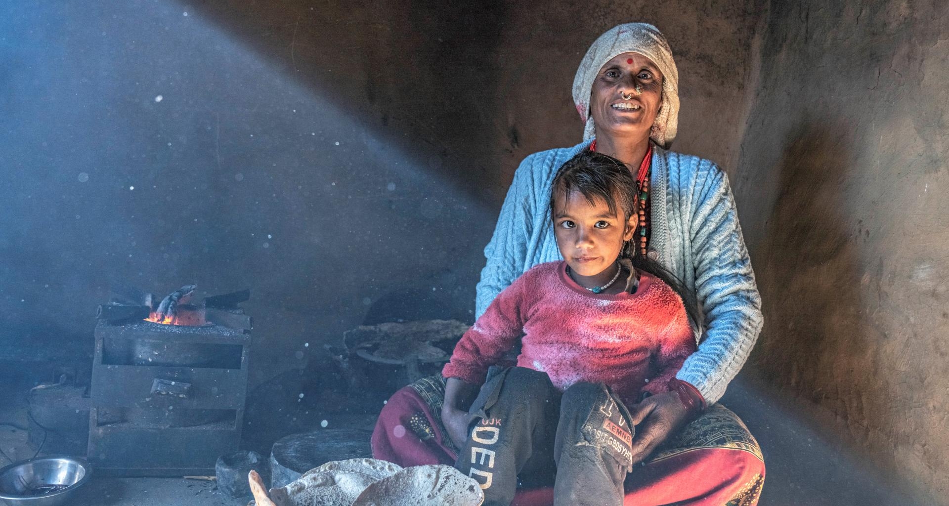 This is a photo clicked on a rural area kitchen where a girl is sitting on her mother's lap with some chapatis in front of her. 